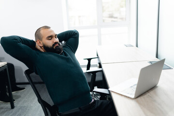 Middle eastern man sitting in contemporary office with relaxed posture, leaning back in his chair near laptop on desk. bright natural light fills space, creating open and airy atmosphere.