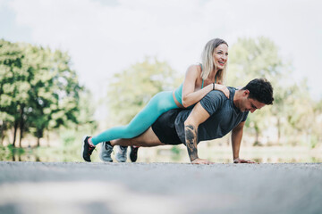 Two friends engage in a playful exercise routine in the park. A man performs push-ups while a woman...