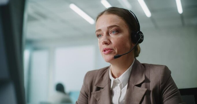 Female marketing manager or customer support specialist works in modern office: Close up of positive Caucasian woman in headset talking to client or business partner, using computer. E-commerce.