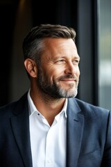 Obraz premium A confident businessman in a sharp suit gazes optimistically out a window in a softly focused setting suggesting contemplation and forward-thinking in modern business.