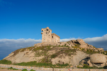 The image shows the ruins of a historic building with a tower and arched windows on a rocky hill in...