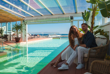 A couple sits by an indoor pool with a glass ceiling, surrounded by tropical plants. Large windows...