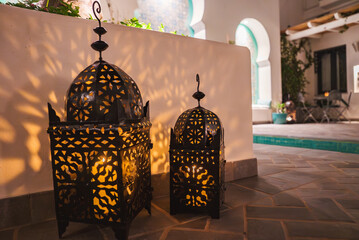 Intricately designed metal lanterns cast shadows on a white wall. A small pool with turquoise water...