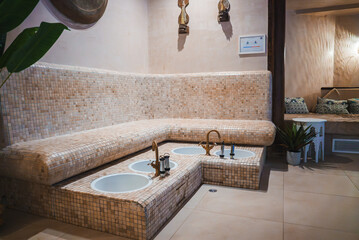 The image shows a lavish interior with mosaic tiles, brass faucets, and foot baths. Hanging...