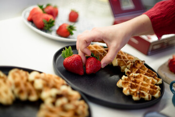 A close-up of a hand carefully placing fresh, vibrant strawberries onto golden waffles on a black plate