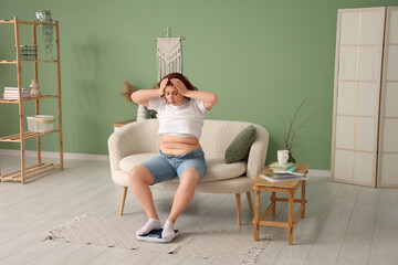 Shocked young overweight woman in tight shorts with scales sitting on sofa at home