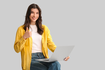 Beautiful young happy woman with laptop sitting on chair and showing thumb-up gesture against on...