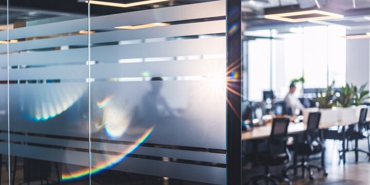 A clear glass partition is in sharp focus, offering a glimpse into a bustling office space with blurred figures working at their desks.