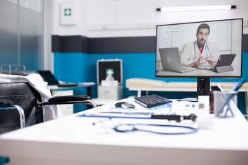 Empty medical room equipped with desktop showing male doctor conducting virtual consultation. Vacant office for wheelchair user reflects the influence of technology on remote health services.
