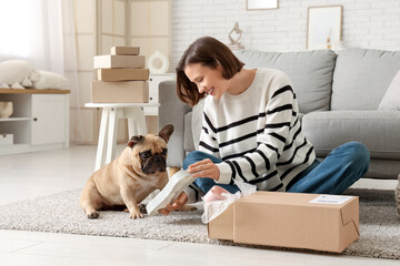 Fototapeta premium Pretty young woman with French bulldog unpacking dog bowl from parcel at home