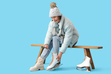 Young woman in winter clothes putting ice skates on bench against blue background