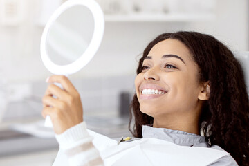 Happy Black Female Patient Looking At Mirror After Dental Treatment In Clinic, Cheerful African...