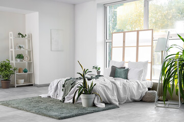 Interior of light bedroom with cozy bed, folding screen and houseplants