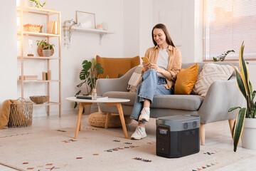 Young woman using mobile phone on sofa with portable power station at home