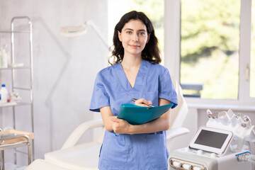 Female cosmetic doctor meticulously stands in her clinic, recording detailed notes in folder,...
