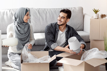 Loving young arab family husband and wife in headscarf unpacking belongings from paper boxes after...