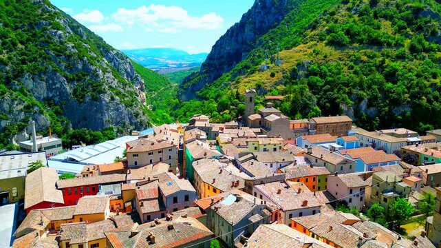 Enchanting aerial push-in fly-over shot from Pioraco, drone flying above the tightly knit buildings and belltower of the town, surrounded by majestic Sibillini Mountains and their green-topped surface