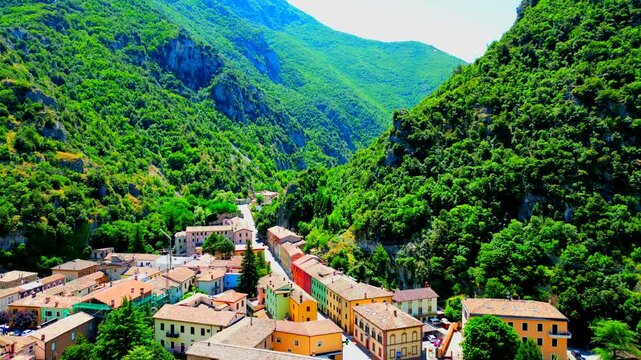 Delicate aerial push-in shot from Pioraco, the sunlit buildings lined at the sides of a road with some cars passing by, perspectivally heading to a gorge deep inside Sibillini mountains' lofty crests