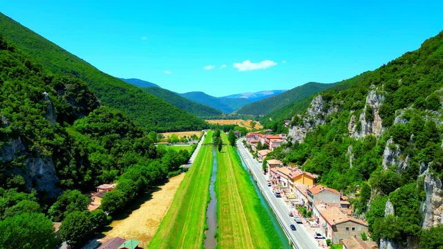 Pristine aerial push-in shot from Pioraco with the Potenza river flanked by gorgeous meadows, road with cars, buildings, and momentous Sibillini mountains with their rocky surface topped by greenery