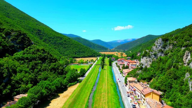 Premium aerial pullback shot from Pioraco with the huge greenery-topped Sibillini Mountains enclosing a valley with the Potenza river, swathes of green shores, road with cars and manifold buildings