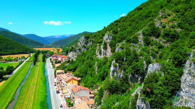 Stunning aerial pan left truck left push-in shot from Pioraco, a close-up of the rocky Sibillini Mountains and their greenery, buildings, roads, the Potenza River flanked by its green shores, cars