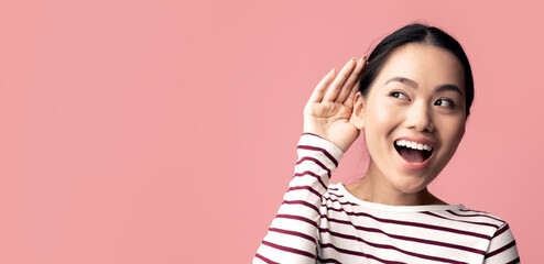Curious cheerful asian woman keeping hand near ear trying to overhear something, interested young...