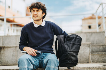 Obraz premium A young student sits on outdoor steps holding a pen, with a backpack beside them. Seems relaxed and thoughtful, perfect for education and lifestyle themes.