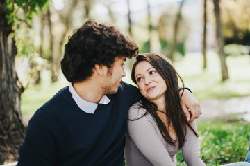 Fototapeta premium A young couple sits closely on a park bench, sharing a tender and affectionate moment. The lush green background adds a serene and romantic atmosphere to their connection.