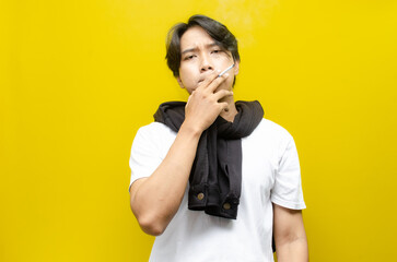 Young Asian Man Smoking a Cigarette and Exhaling Smoke Isolated on Yellow Background