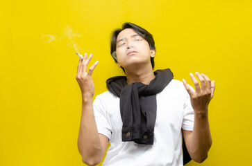 Young Asian Man Smoking a Cigarette and Exhaling Smoke Isolated on Yellow Background