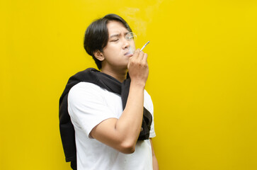 Young Asian Man Smoking a Cigarette and Exhaling Smoke Isolated on Yellow Background