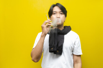 Young Asian Man Smoking a Cigarette and Exhaling Smoke Isolated on Yellow Background