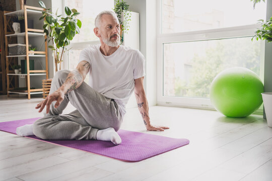 Elderly man practicing yoga on mat in bright room with natural light and green exercise ball nearby