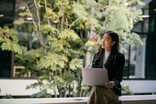 Young businesswoman sits with her laptop in a modern office space adorned with lush greenery, looking thoughtfully towards upcoming projects and opportunities