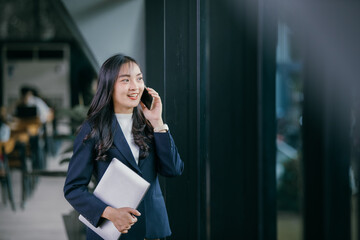 Smiling young businesswoman talking on the phone while holding a laptop, standing confidently in a...