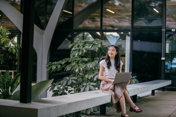 Young asian freelancer is sitting on a bench, working on her laptop in the outdoor area of a modern...