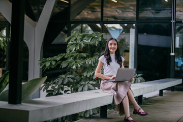 Young asian woman using laptop while sitting on bench in modern office space with green plants, working remotely as freelancer or digital nomad enjoying flexible work environment - Powered by Adobe