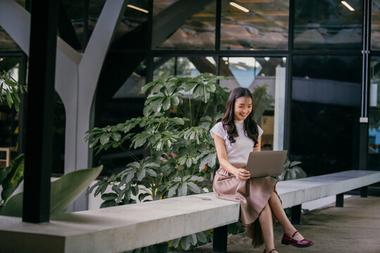 Young asian freelancer is smiling while working remotely on her laptop, enjoying the modern and green office space around her, promoting a productive and pleasant work environment
