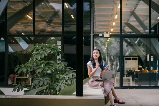 Young asian businesswoman is sitting on a bench outside a modern office building, using her laptop and smartphone while enjoying the benefits of a flexible work arrangement