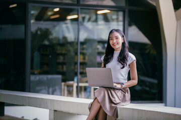 Young asian woman working as freelancer using laptop, sitting on a wall outside a modern building...