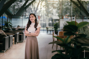 Portrait of a smiling young asian businesswoman standing with arms crossed in a modern office lobby, showcasing confidence and professionalism in a contemporary workspace