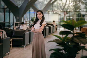 Portrait of a smiling young asian businesswoman standing with arms crossed in a modern office lobby, exuding confidence and professionalism in her stylish attire