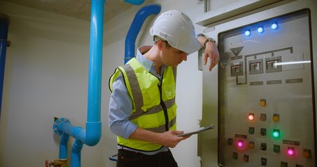 A technician Engineer in a safety vest and helmet inspects a control panel in Industrial Facility, using a Digital Tablet to monitor Maintenance machinery lights and system status in an industrial