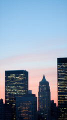 Obraz premium Phone wallpaper of a city skyline with glowing lights in the buildings beneath a soft, gradient sky transitioning from warm pink hues to cool blue tones at dusk