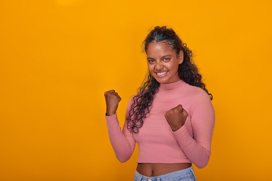 Young Indian woman stands confidently with her fists raised in triumph. Her bright smile complements the vibrant yellow background, radiating positivity and energy in a playful atmosphere.