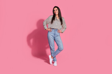 Young woman standing confidently on pink background in casual striped shirt and jeans, exuding...