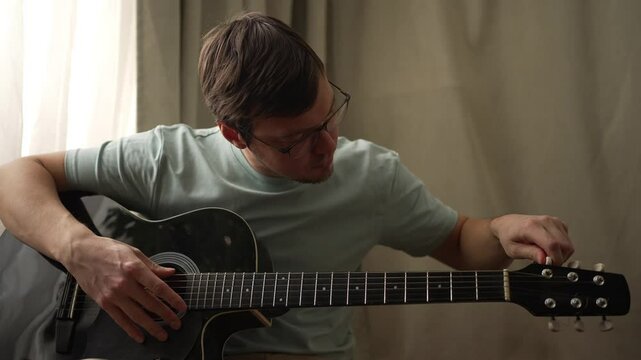 Man in casual clothes tunes the strings of an acoustic guitar with care. Adjusting each tuning peg one by one, he focuses on achieving the perfect sound quality