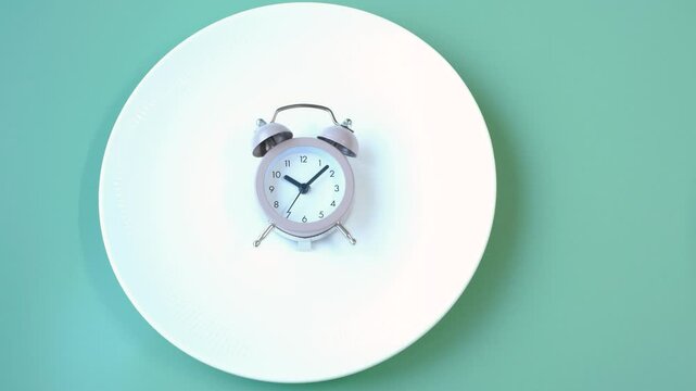 Female hand holding alarm clock against empty plate, intermittent fasting concept.