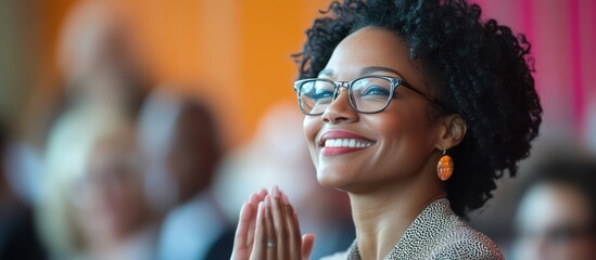 Fototapeta premium A woman with curly hair and glasses smiles while clasping her hands together.
