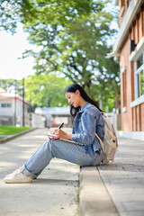 A positive Asian female student sits on the edge of a footpath at her university, writing in a book.